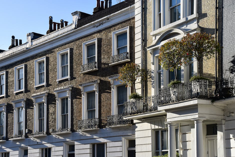 Image of a traditional multi-story residential building with brick facades and white window frames, featuring small decorative balconies with wrought iron railings. The buildings are illuminated by sunlight, casting shadows on the facade. The windows are clean, with some open and others closed, and there are potted plants on the balconies adding greenery. Visible are elements of the building's exterior including cornices, window sills, and a section of the entranceway. The overall appearance reflects well-maintained, classic urban architecture, emphasizing the importance of regular surface cleaning and maintenance for historic buildings, as promoted by Cleaner Fulham in their Fulham Road flat cleaning guide for residents.