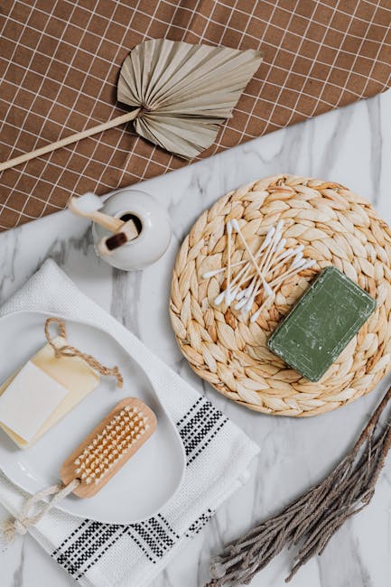 A flat lay arrangement of bathroom or spa accessories on a white marble surface, including a round woven placemat with a green soap bar and a pile of cotton swabs, a wooden brush with bristles, a white ceramic dispenser, and a lemon-colored towel. The scene features natural tones with beige, white, and green accents, illuminated by soft, natural lighting. A dried palm leaf lies nearby, adding a touch of organic texture. This image showcases elements related to personal hygiene and surface cleaning, reflecting the focus of domestic cleaning and sanitisation services offered by Cleaner Fulham, as referenced on the Parsons Green end of tenancy cleaning tips page.